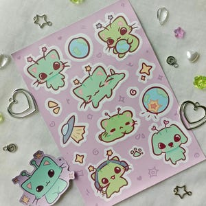 May include: A sheet of stickers featuring cartoon alien cats, planets, and a UFO, in shades of green, pink, and blue. The stickers have a cute, whimsical design, with stars and hearts. The stickers are on a pink background.