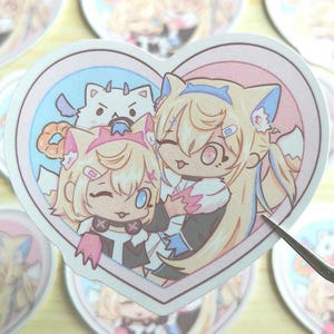 May include: A heart-shaped sticker with two anime-style characters. The characters are both female with blonde hair and wearing cat ears. One character is wearing a black and white outfit and the other is wearing a white and blue outfit. The sticker is on a light brown wooden surface.