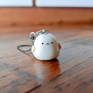 May include: A white ceramic bird charm with black eyes and a small black string attached. The charm is shaped like a bird with a round body and a small beak.