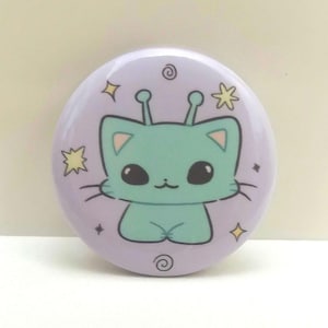 Puede incluir: Un botón morado con un gato alienígena verde de dibujos animados con antenas. El gato tiene ojos grandes y una expresión linda. El fondo es morado claro con estrellas amarillas y remolinos.