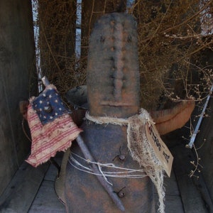 Pu&ograve; includere: Una bambola di pezza rustica, marrone e fatta a mano, con una sciarpa di iuta e una piccola bandiera americana. La bambola tiene un bastone e ha un viso cucito.