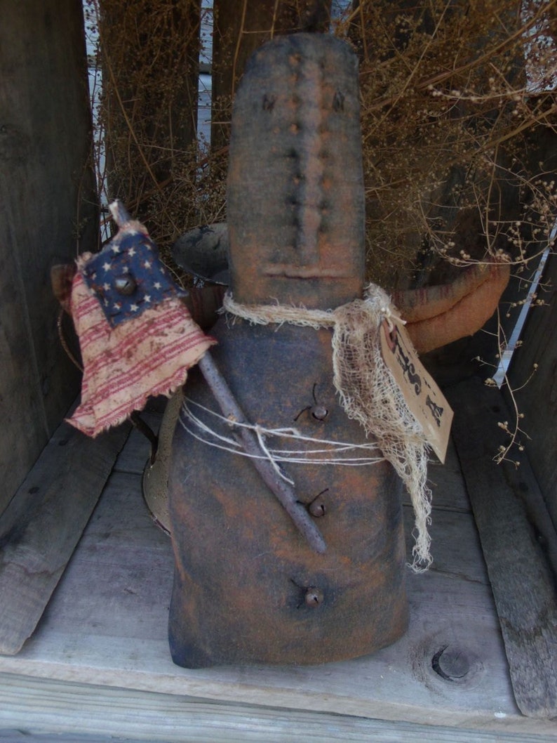 Pu&ograve; includere: Una bambola di pezza rustica, marrone e fatta a mano, con una sciarpa di iuta e una piccola bandiera americana. La bambola tiene un bastone e ha un viso cucito.