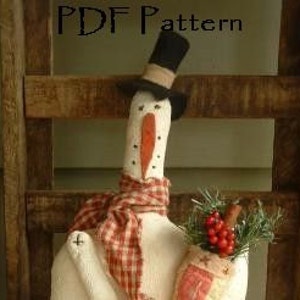 May include: A white fabric snowman with a red and white plaid scarf, a black top hat, and a red nose. The snowman is holding a small burlap bag with a red and white design and a sprig of greenery with red berries.