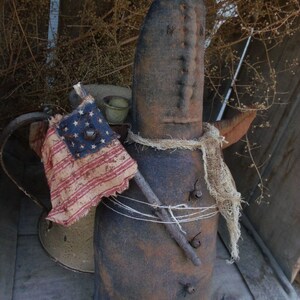 Pu&ograve; includere: Una bambola di pezza rustica fatta a mano con un corpo in tessuto marrone e una sciarpa di iuta. La bambola tiene un bastone con una piccola bandiera americana attaccata. La bambola &egrave; seduta su una superficie di legno.