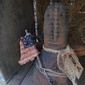 Pu&ograve; includere: Una bambola di pezza rustica fatta a mano con un corpo di sacco di iuta e un viso invecchiato. La bambola tiene una piccola bandiera americana e ha una sciarpa di iuta bianca legata intorno al collo. La bambola &egrave; seduta su una superficie di legno con un'etichetta che dice "Tess A".