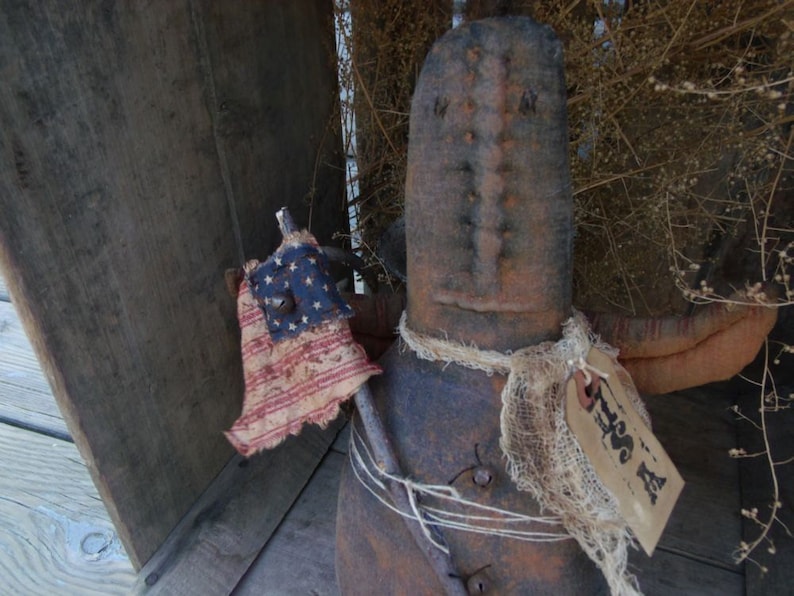 Pu&ograve; includere: Una bambola di pezza rustica fatta a mano con un corpo di sacco di iuta e un viso invecchiato. La bambola tiene una piccola bandiera americana e ha una sciarpa di iuta bianca legata intorno al collo. La bambola &egrave; seduta su una superficie di legno con un'etichetta che dice "Tess A".