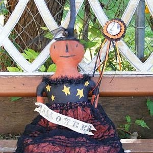 May include: A handmade rag doll dressed as a witch for Halloween. She is wearing a black dress with orange lace trim and a black hat with a star design. She is holding a wand with a star on top. The doll is sitting on a wooden bench with a white lattice background.