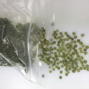 May include: A plastic bag filled with small, green, faceted gemstones. Some of the gemstones have spilled out of the bag onto a white surface.