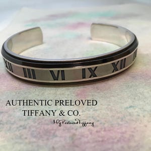 Puede incluir: Brazalete de plata con detalles negros y números romanos. El brazalete lleva la inscripción "AUTHENTIC PRELOVED TIFFANY & CO." y "My Preloved Tiffany."