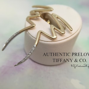 May include: Gold-tone earrings with a zigzag design, featuring a row of small diamonds on the curved ends. The earrings are displayed on a white pedestal, with the text "AUTHENTIC PRELOVED TIFFANY & CO."
