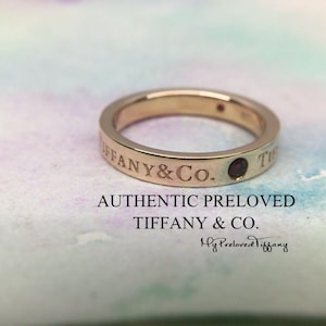Può includere: Un anello a fascia d'oro con l'iscrizione "Tiffany & Co." e una piccola pietra rossa scura. L'anello è su uno sfondo bianco e rosa.