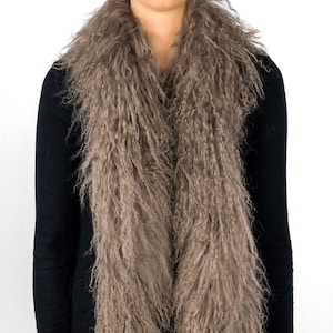 Może przedstawiać: Długi, brązowy szal z imitacji futra o kręconej fakturze. Szal jest noszony wokół szyi osoby w czarnej koszuli z długim rękawem i niebieskich dżinsach.