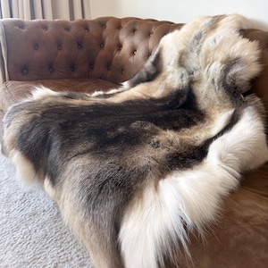 May include: A luxurious, natural fur throw draped over a brown leather sofa. The throw features a mix of white, brown, and black fur, creating a striking contrast. The sofa has a tufted design, adding to the elegant aesthetic. An abstract painting hangs on the wall.