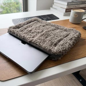 May include: A grey laptop partially inserted into a rectangular, taupe-colored, fuzzy, sherpa-style fleece sleeve. The sleeve features a thick, textured, plush exterior with a zippered closure along the top edge, designed to protect the electronic device.