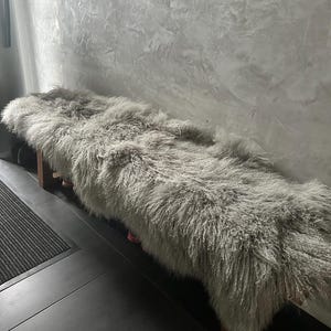 Puede incluir: Una alfombra de piel de oveja gris larga y esponjosa se coloca sobre un banco de madera. La alfombra es suave y mullida, y ser&iacute;a perfecta para agregar un toque de calidez y comodidad a cualquier habitaci&oacute;n.