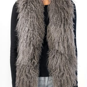Peut inclure: Un gilet en fourrure gris et duveteux avec une longue texture hirsute. Le gilet est porté sur un haut noir à manches longues et un jean gris clair. La fourrure semble être de couleur naturelle et le gilet est ouvert à l'avant.