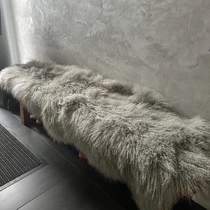 Puede incluir: Una alfombra de piel de oveja esponjosa y gris claro se coloca sobre un banco de madera. La alfombra es suave y mullida, con fibras largas y fluidas. El banco es de madera oscura y tiene un dise&ntilde;o sencillo.