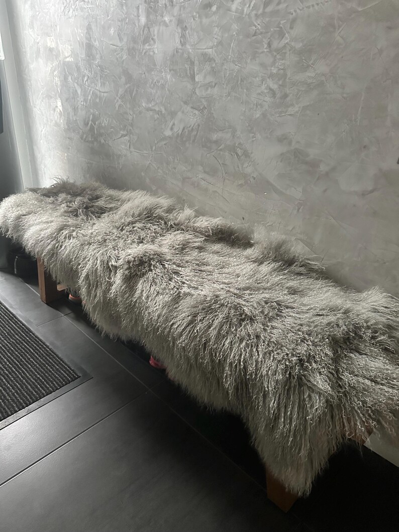 Puede incluir: Una alfombra de piel de oveja esponjosa y gris claro se coloca sobre un banco de madera. La alfombra es suave y mullida, con fibras largas y fluidas. El banco es de madera oscura y tiene un dise&ntilde;o sencillo.
