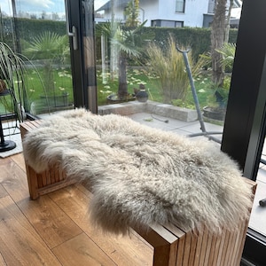 May include: A light gray sheepskin rug placed on a wooden bench with slatted sides. The bench is in a room with a wooden floor and a large window behind it.