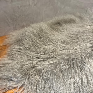 Puede incluir: Una alfombra de piel de oveja gris y esponjosa con fibras largas y rizadas. La alfombra est&aacute; colocada sobre una superficie de madera.