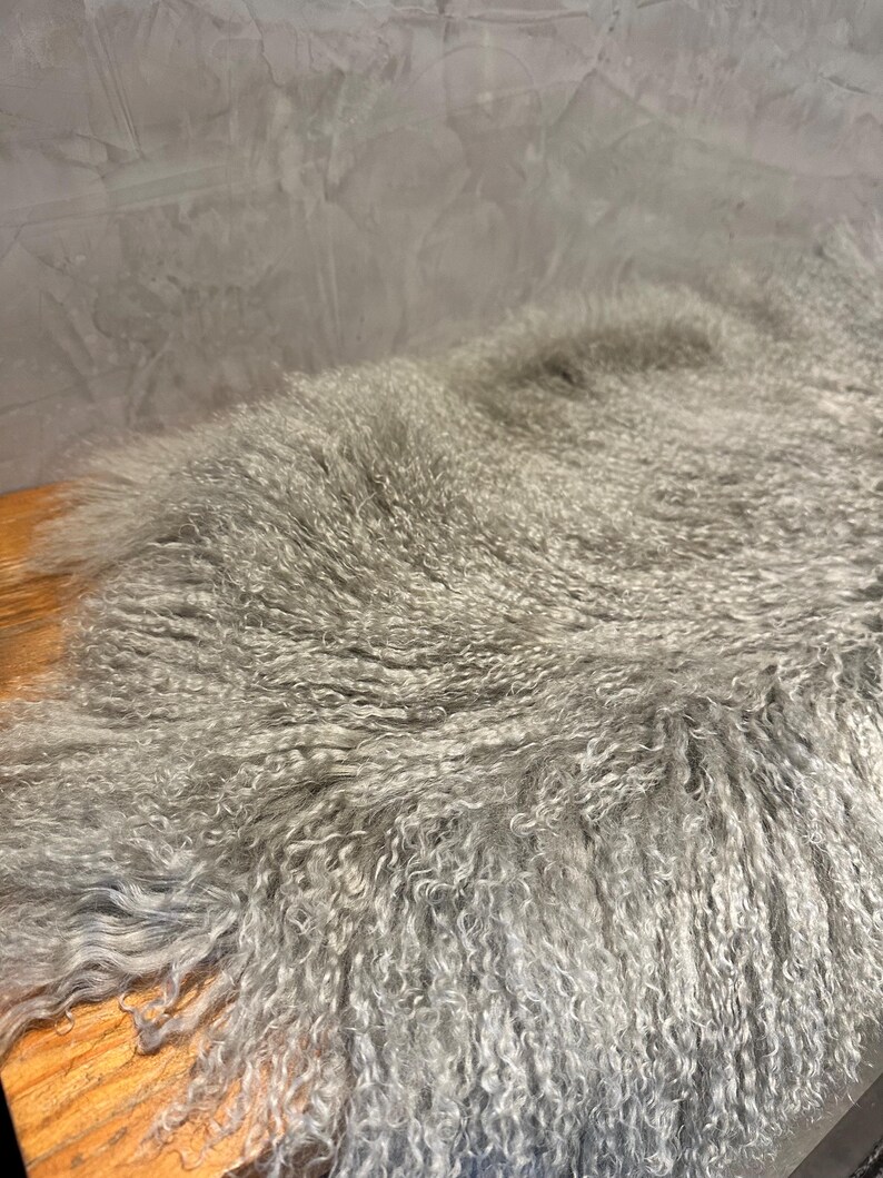 Puede incluir: Una alfombra de piel de oveja gris y esponjosa con fibras largas y rizadas. La alfombra est&aacute; colocada sobre una superficie de madera.