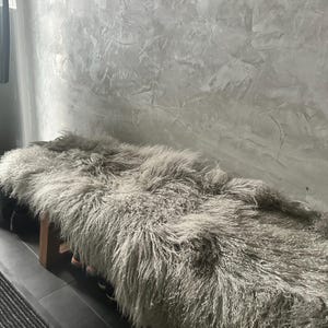 Puede incluir: Una alfombra de piel de oveja gris esponjosa est&aacute; drapeada sobre un banco de madera. La alfombra es suave y mullida, y el banco est&aacute; hecho de madera natural.