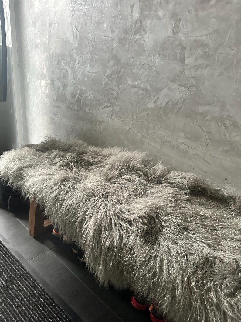 Puede incluir: Una alfombra de piel de oveja gris esponjosa est&aacute; drapeada sobre un banco de madera. La alfombra es suave y mullida, y el banco est&aacute; hecho de madera natural.
