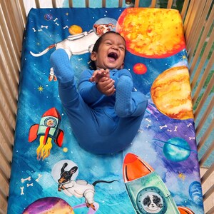 May include: A blue crib sheet with a space-themed design featuring planets, stars, and rockets. Two dogs wearing astronaut suits are flying in space. One dog is in a rocket and the other is flying through the air. There are white bones scattered throughout the design.