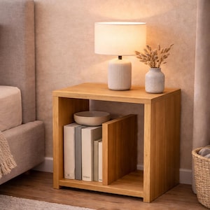 Puede incluir: Una mesita de noche de roble claro con una lámpara, un jarrón y libros. La lámpara tiene una pantalla y una base blancas. Un pequeño jarrón con flores secas está junto a la lámpara. La mesita mide 61 cm de ancho.