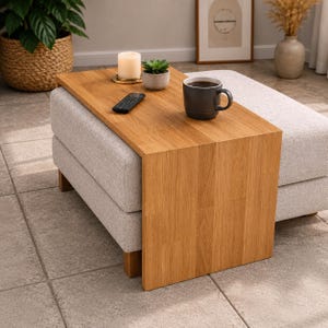 Könnte beinhalten: Ein hell gepolsterter Hocker mit einem hölzernen Beistelltisch. Der Tisch aus hellem Holz trägt eine Kerze, eine kleine Pflanze, eine Fernbedienung und eine Tasse. Der Hocker ist mit hellgrauem Stoff bezogen.