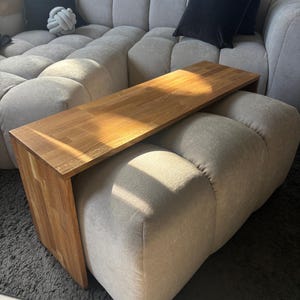 Peut inclure: Une table d'appoint rectangulaire en bois avec une finition brun clair et un grain de bois visible, dotée d'une surface supérieure plate et d'un panneau latéral vertical unique qui descend jusqu'au sol, posée sur un pouf rembourré clair.