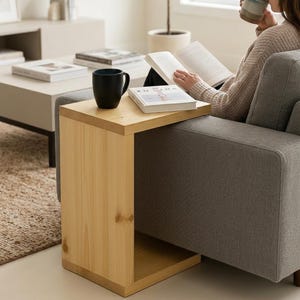 Könnte beinhalten: Ein helles Holz-Beistelltisch mit rechteckiger Oberseite und quadratischer Basis. Ein schwarzer Becher und ein Buch liegen auf dem Tisch. Der Tisch steht neben einem grauen Sofa und schafft eine gemütliche Leseecke.