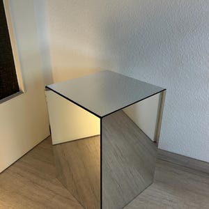 Puede incluir: Una mesa auxiliar con forma de cubo espejado y borde negro. Las superficies reflectantes crean una estética moderna y minimalista. La mesa está colocada sobre un suelo de madera.