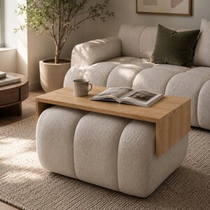 May include: A light beige upholstered ottoman with a rounded, segmented design featuring a rectangular light wood tray table placed across the top. An open book and a ceramic mug are positioned on the wooden surface of the tray table.