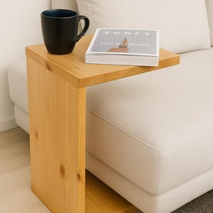 Könnte beinhalten: Kleiner, hellfarbiger Beistelltisch aus Holz mit rechteckiger Platte und vertikaler Stütze. Eine schwarze Tasse mit blauem Innenraum und ein Buch mit dem Text "ZUNFT ZUR OFFNUNG" stehen auf dem Tisch. Der Text "Kiefer Geölt" steht oben links.