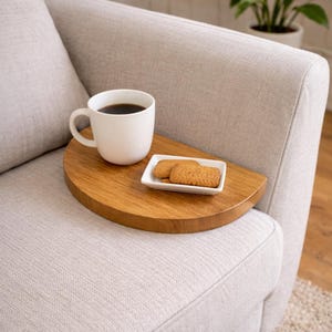 May include: A wooden half-circle tray holds a white coffee mug with dark liquid and a small white plate with two biscuits. The tray rests on the arm of a light grey upholstered sofa. A plant in a white pot is in the background.