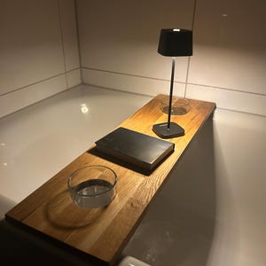 Puede incluir: Una bandeja de baño de madera con una lámpara negra, un libro y una taza de vidrio. La bandeja está colocada en el borde de una bañera blanca.