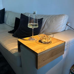May include: A wooden sofa arm table with a glass of white wine and a bowl of nuts. The table is made of light-coloured wood and is attached to the arm of a white leather sofa. Black cushions are visible.