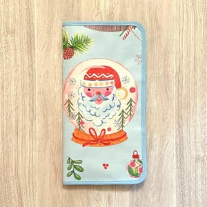 May include: A light blue Christmas-themed zippered case featuring a snow globe with Santa Claus. The case has a light blue border and a festive design with pine needles, snowflakes, and a Christmas ornament.