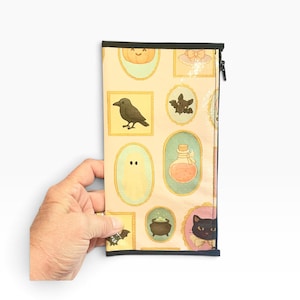 May include: A rectangular zippered pouch with a cream-colored background featuring a repeating pattern of framed illustrations including a pumpkin, a black crow, a bat, a ghost, a potion bottle, a cauldron, and a black cat with a bow tie.