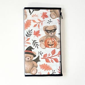 May include: A rectangular zippered pouch featuring a white background with a repeating pattern of brown teddy bears, orange pumpkins, and autumn leaves. One bear wears a black mask, while another wears a black witch hat and bat wings.