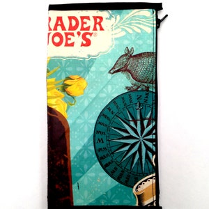 Op de afbeelding: Een rechthoekig Trader Joe's etui met ritssluiting met een turquoise achtergrond met een kompas, een gordeldier en een gele bloem. Het Trader Joe's-logo staat bovenaan in rode letters.