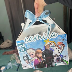 Puede incluir: Una caja de regalo azul y blanca con un lazo azul y un tema de Frozen. La caja tiene un asa y está decorada con imágenes de Elsa, Anna, Olaf y Kristoff. La caja también tiene el nombre "camilla" y el número "3" en ella.