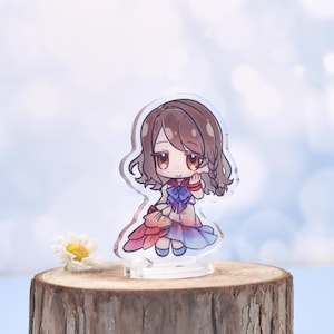May include: A clear acrylic anime-style figure of a girl with brown hair and a colorful dress. The figure is standing on a clear base and is placed on a wooden surface. The background is a soft blue with white bokeh.