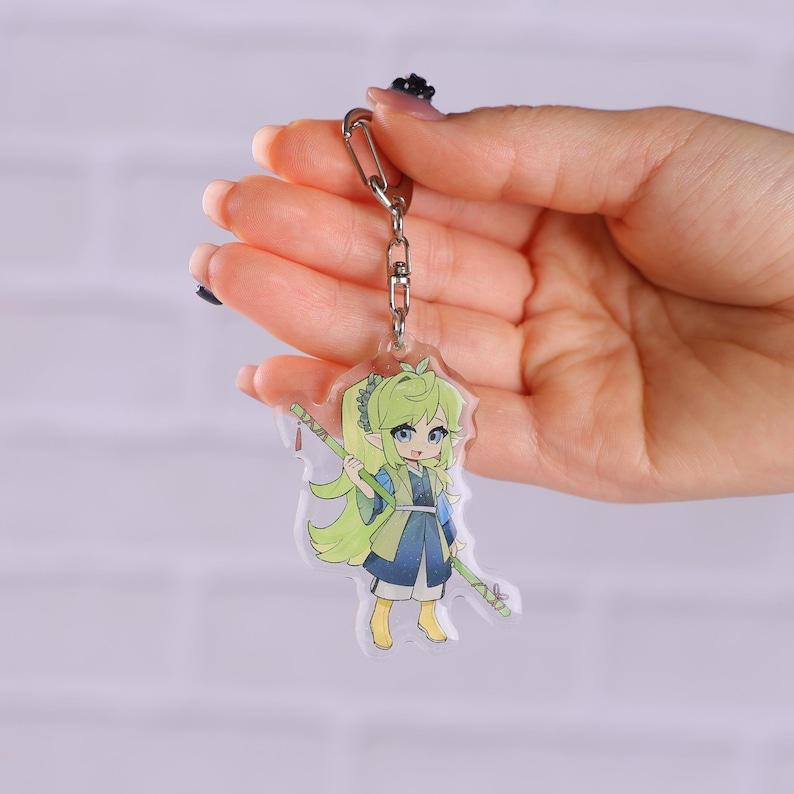 May include: A clear acrylic keychain featuring a cartoon character with green hair, blue eyes, and a blue and yellow outfit. The character is holding two green staffs. The keychain has a silver metal clasp.