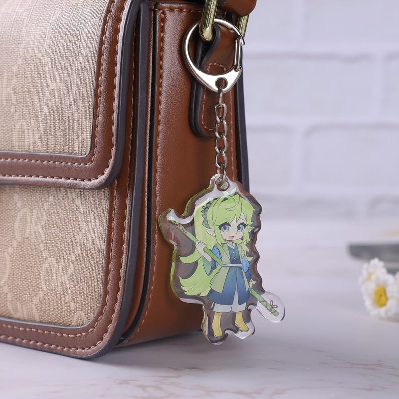 May include: A clear acrylic charm featuring a cartoon character with green hair and blue eyes, holding a bamboo staff. The charm is attached to a silver keychain, hanging from a brown and tan handbag.