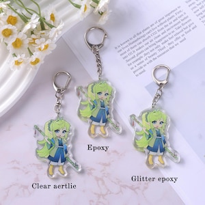 May include: Clear acrylic keychains featuring a cartoon anime girl with green hair and a blue and white outfit, holding a staff. The keychains are attached to silver keyrings. One keychain is labeled "Epoxy" and another "Glitter epoxy".