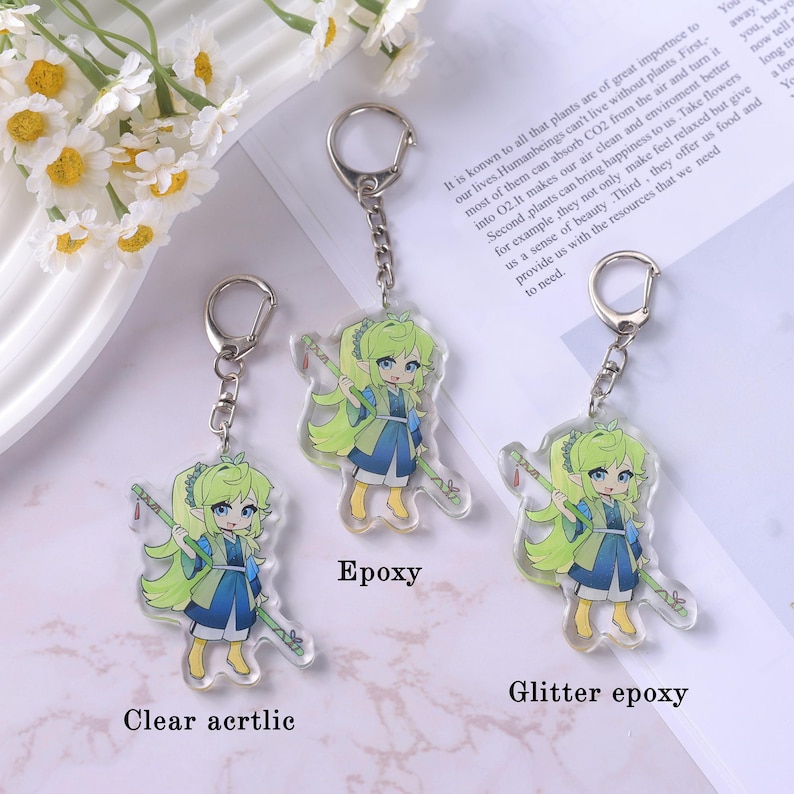 May include: Clear acrylic keychains featuring a cartoon anime girl with green hair and a blue and white outfit, holding a staff. The keychains are attached to silver keyrings. One keychain is labeled "Epoxy" and another "Glitter epoxy".