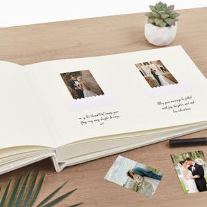 May include: An open, off-white wedding photo album with two photos displayed. The album has handwritten text on the pages. Additional photos are scattered on the wooden surface. A small succulent plant in a white pot is in the background.