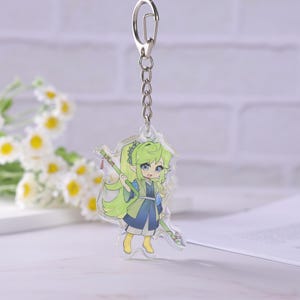 May include: A clear acrylic keychain featuring a cartoon character with green hair and blue eyes, holding a green staff. The character is wearing a blue and green outfit. The keychain has a silver metal chain and clasp.